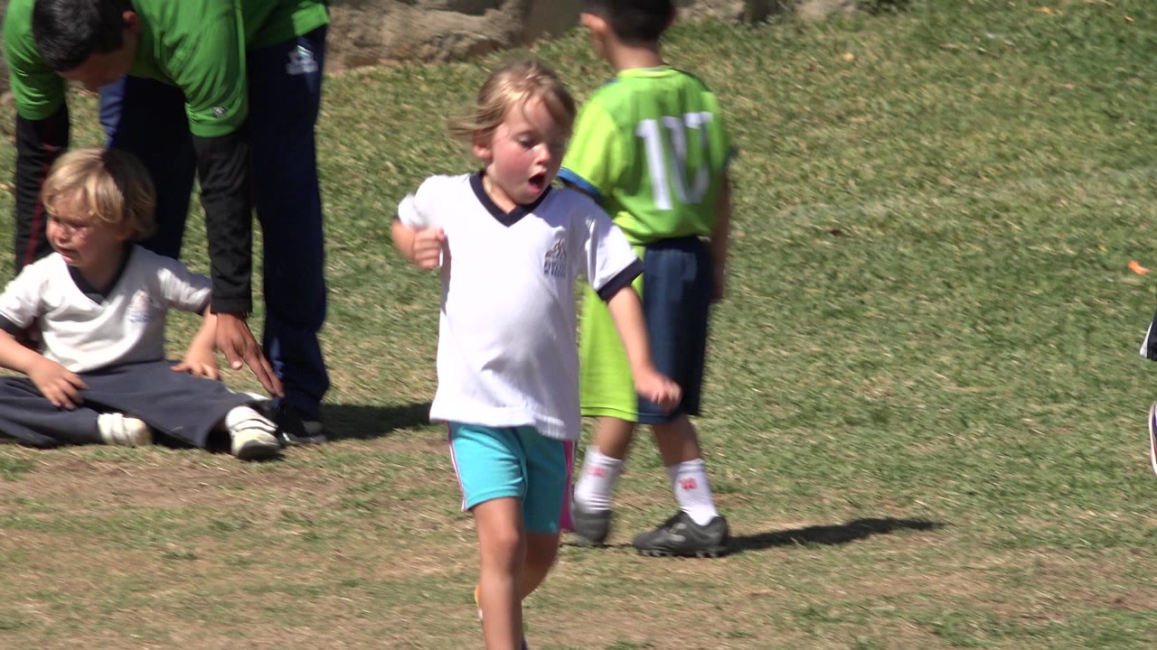 One second in, the blond child remains near the foreground as a child in a green jersey and a seated child appear behind on the grassy field in San Jose Del Cabo.