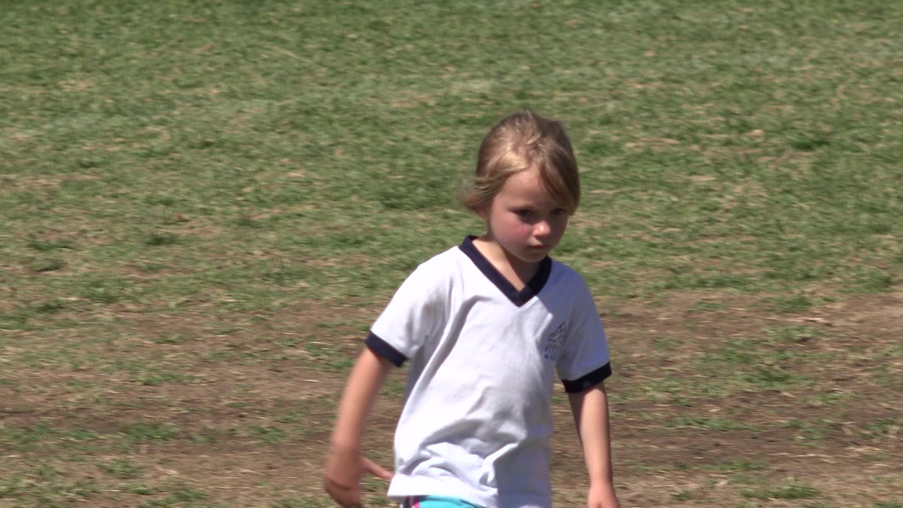 A blond child in a white sports jersey walks across a sunlit grass field in San Jose Del Cabo, Mexico, with the body centered and no ball visible.