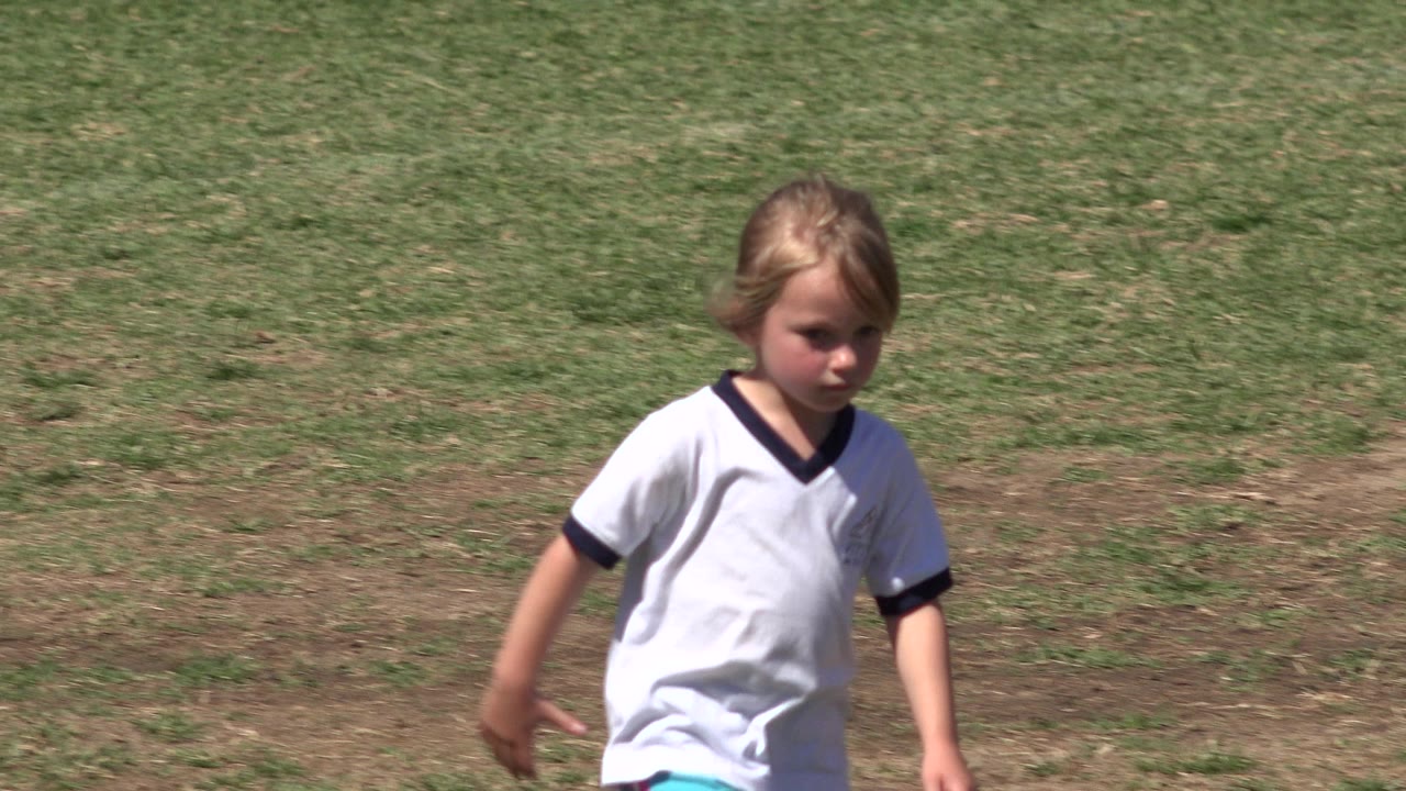 The child in the white jersey continues walking through the sunlit San Jose Del Cabo grass field, with the face turned toward the camera and the background mostly open grass.