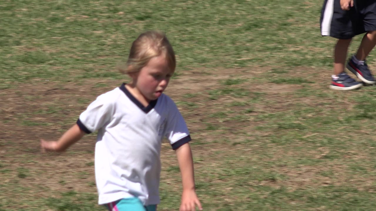Young child walking on grass with cropped players nearby.