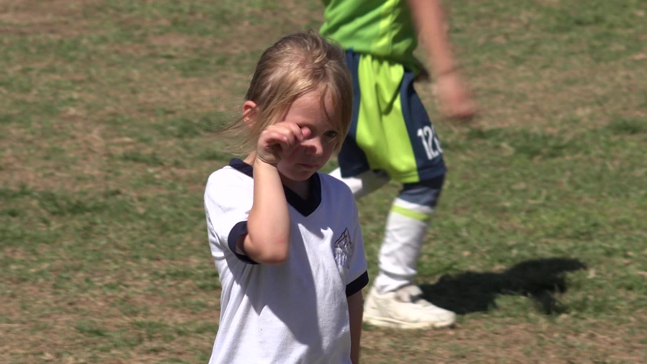 Young girl in a white soccer jersey on a grass field with a player behind her.
