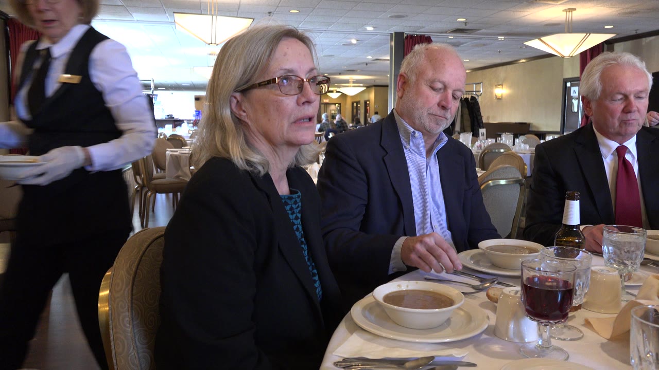In a Chicago restaurant, three business-dressed adults sit at a lunch table as a server passes close to the camera with bowls, with soup, glasses, and beer bottles on the table.