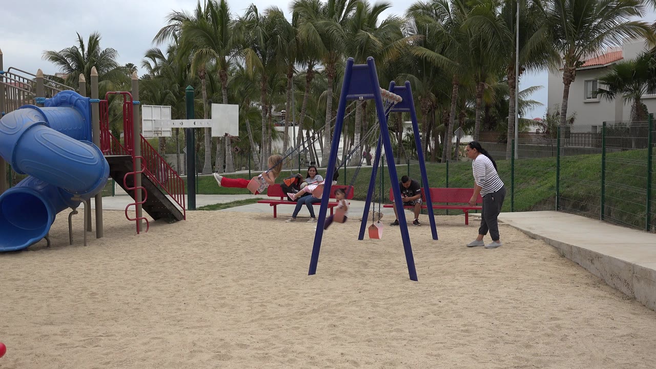 Wide view of a San Jose del Cabo playground where children play near swings as adults sit and stand by a red bench, with a blue tube slide and palm trees behind them.
