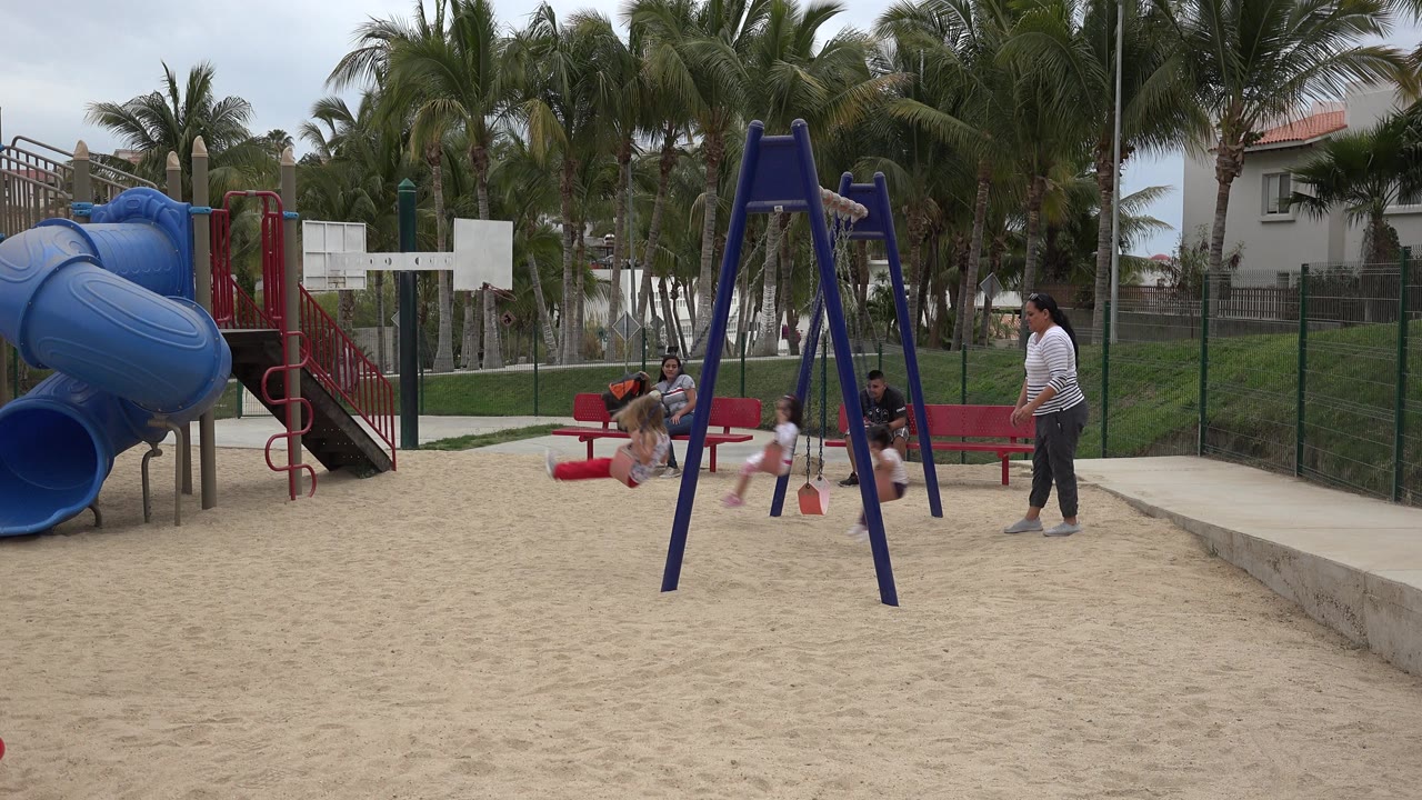 A child is near the swing set at the San Jose del Cabo playground while adults sit on red benches and another adult stands beside the sand area.