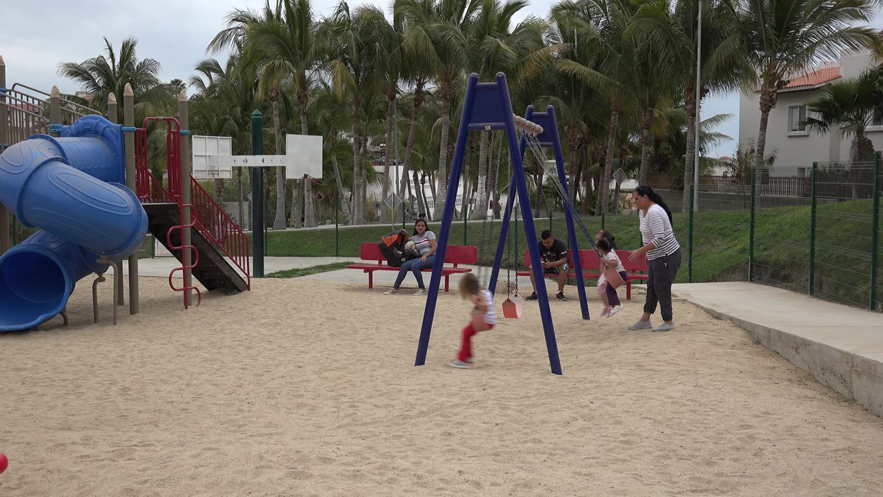 Children and adults are visible at a sandy playground in San Jose del Cabo, Mexico, with a blue spiral slide on the left, swings in the center, red benches behind, and palm trees in the background.