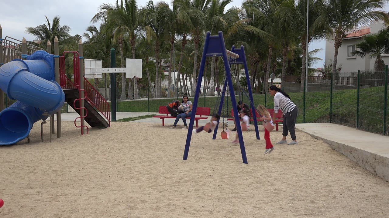 Children use the swings while adults watch from the bench and walkway.