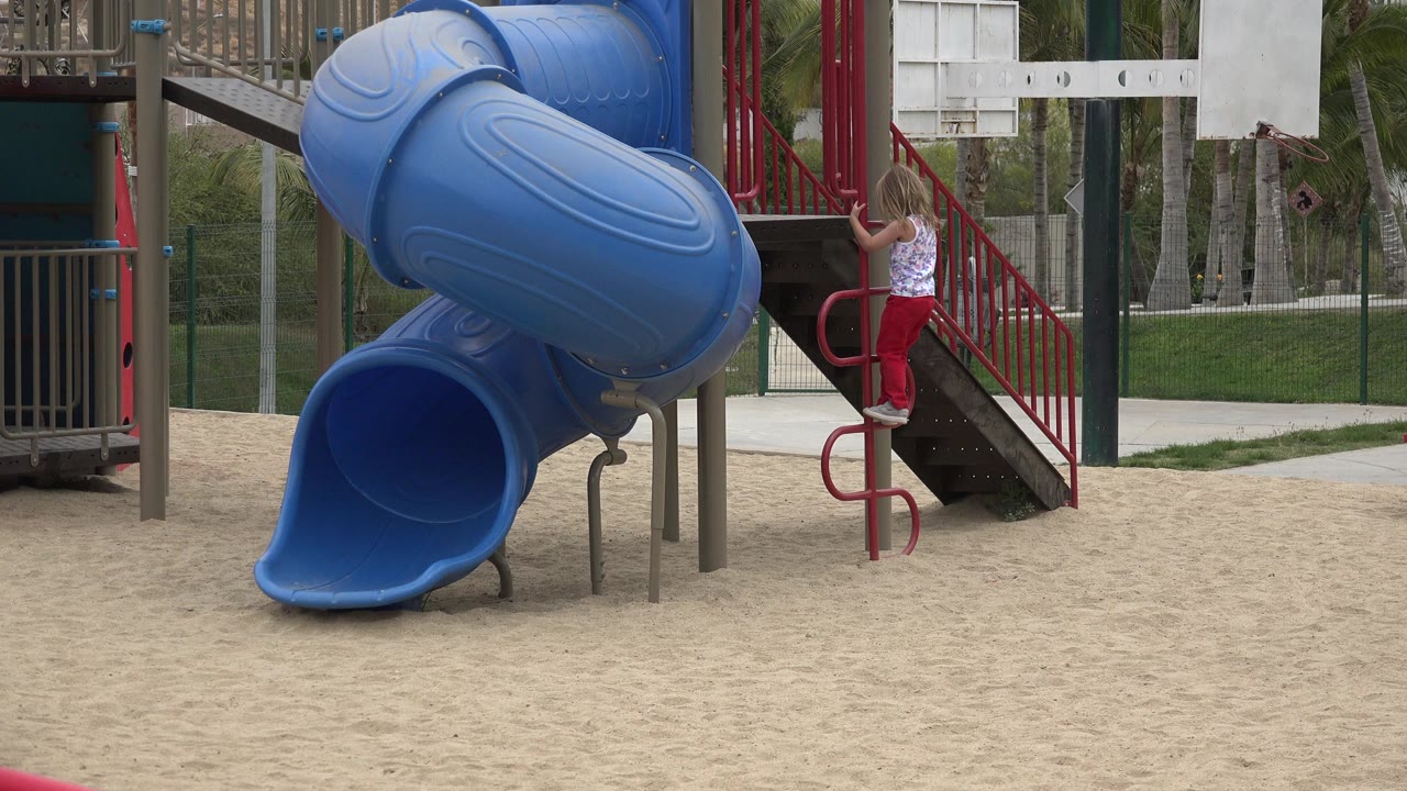 The girl is slightly higher on the red ladder, holding the rails while the blue tube slide fills the left side of the San Jose Del Cabo frame.