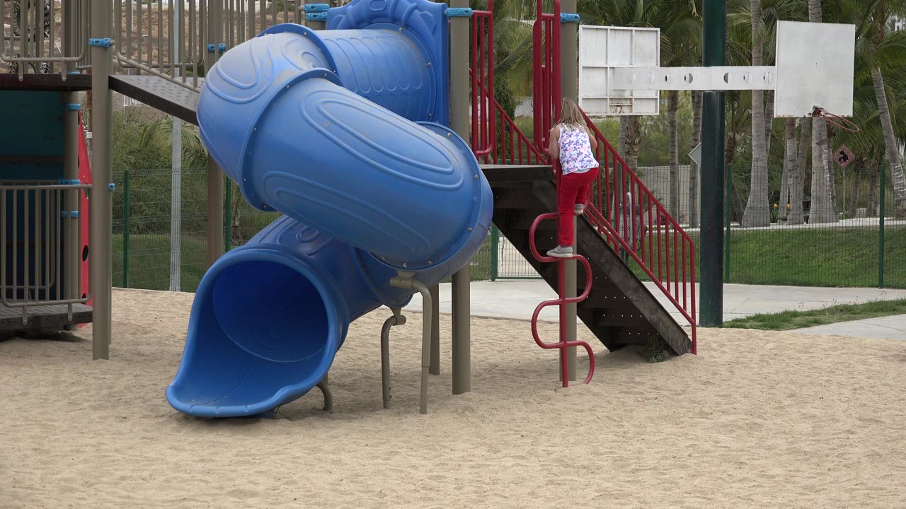 Blonde girl climbing a red playground ladder beside a blue tube slide.