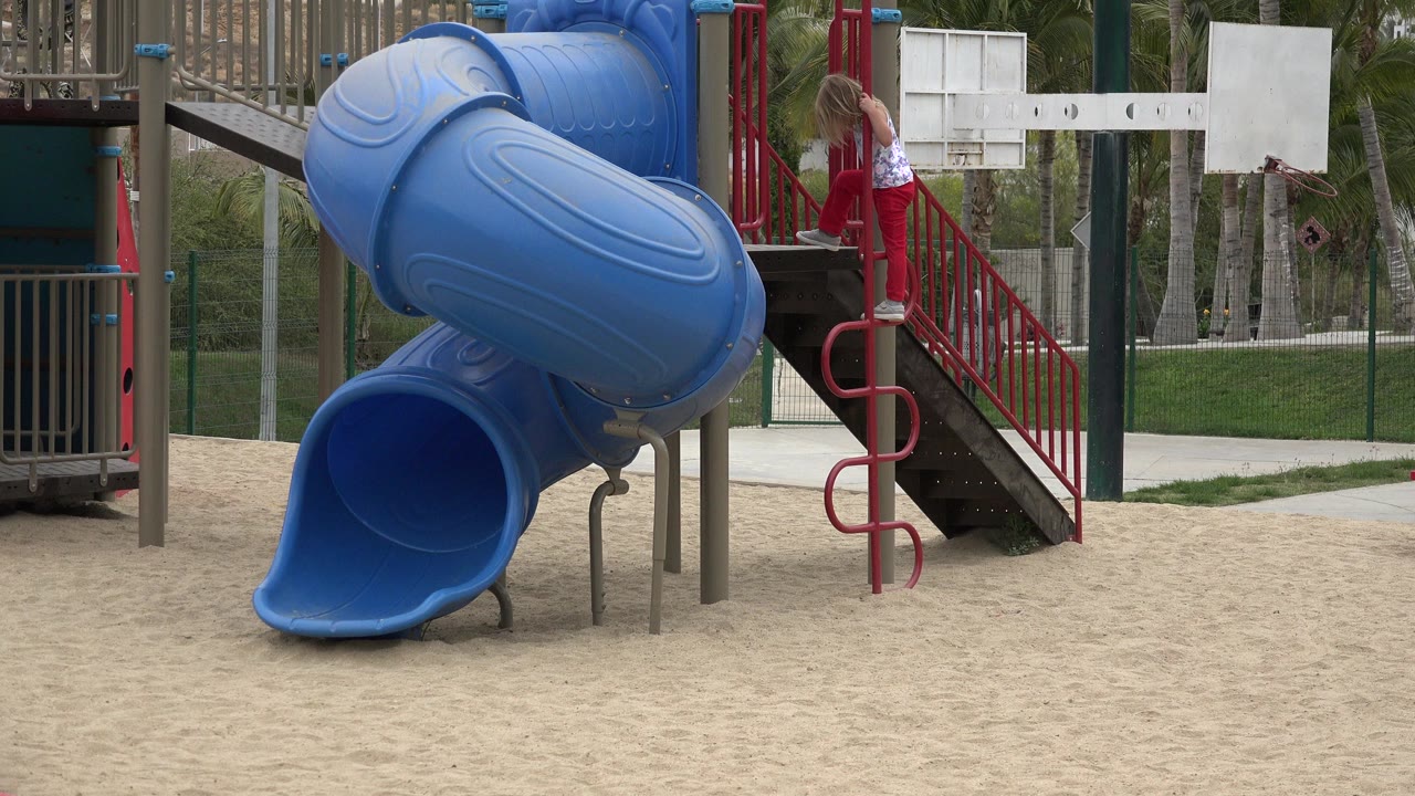 A child in red pants climbs near a red spiral ladder beside a large blue playground slide in San Jose del Cabo.