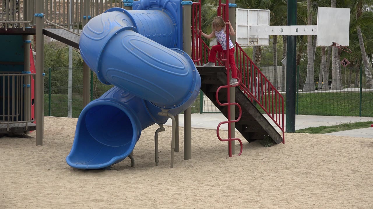 At 0:02 in San Jose del Cabo, Mexico, the blond child remains on the right platform area beside red rails, with the rounded blue slide body in the foreground.