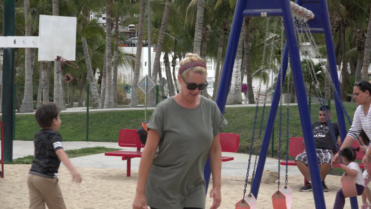 Woman in sunglasses walking near playground swings with children and adults behind her.