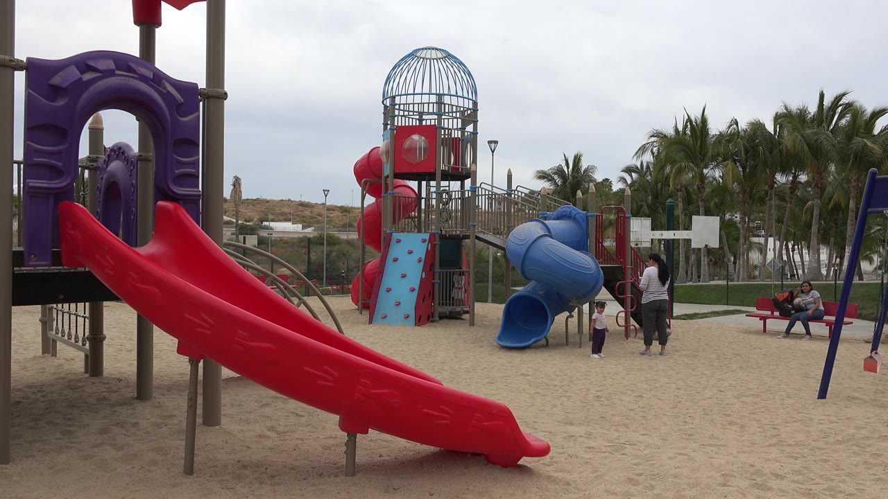 Sandy playground with a red slide, blue tube slide, and people near the play structure.