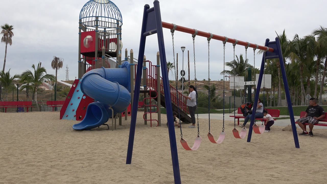 Sandy playground with swings, slide structure, palm trees, and people on benches.