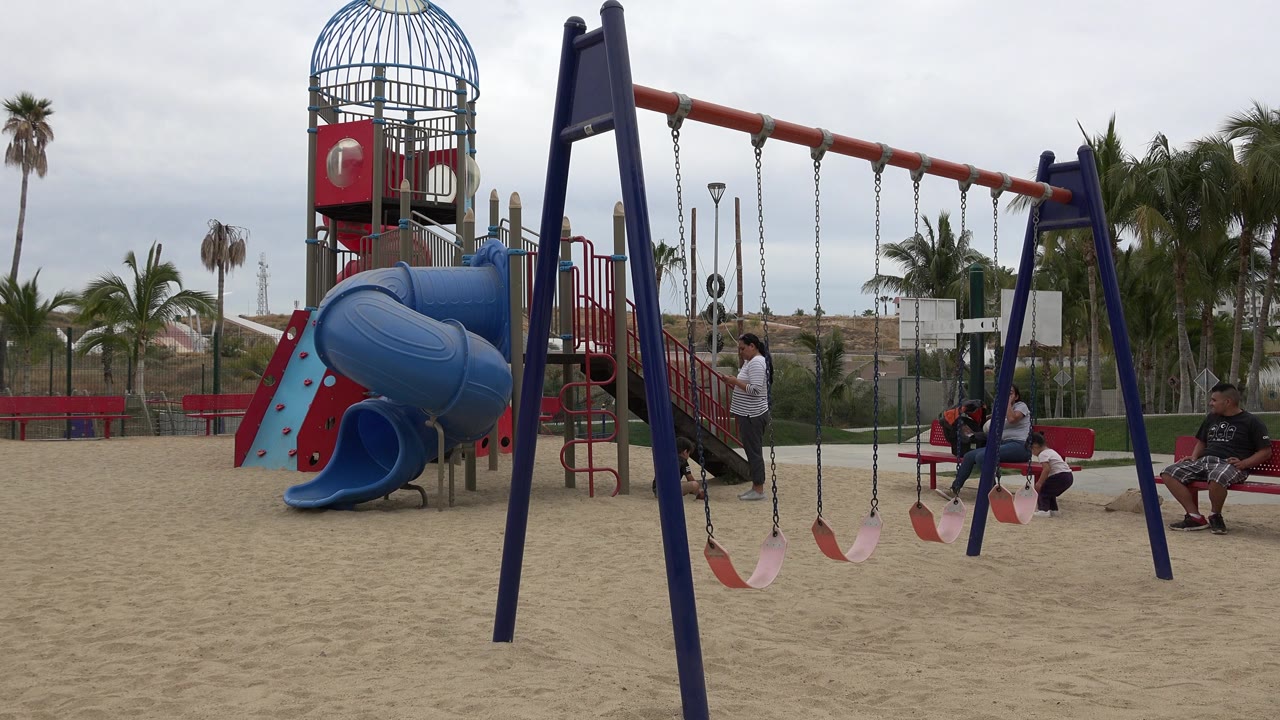 San Jose del Cabo playground view with empty swing seats hanging in the foreground, a person standing by the play structure, and seated people near red benches.