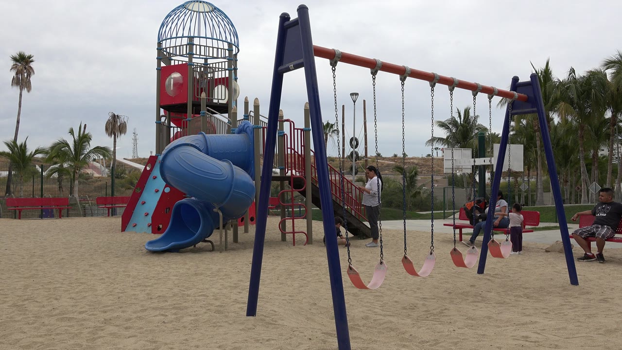 Sandy playground with swing set, slide tower, palms, and people near benches.