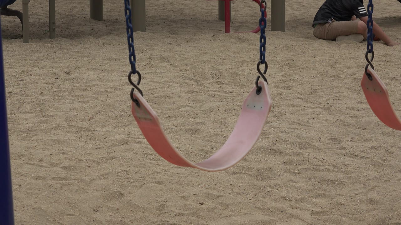 An empty pink and red swing hangs low over sand in a San Jose del Cabo playground, with blue chains in the foreground and a partial seated person visible near the top edge.