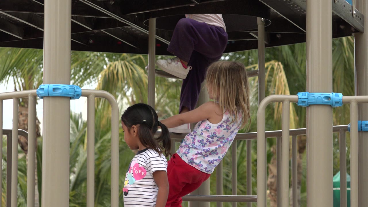 Children climb and stand near tan playground railings in San Jose del Cabo, Mexico, with palm trees visible beyond the structure.