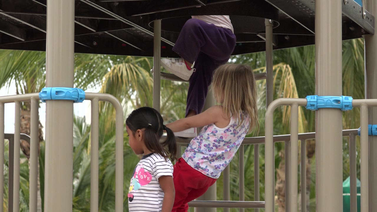 Two children remain beside the tan playground railings in San Jose del Cabo, Mexico, while a third child is partly visible climbing above them.