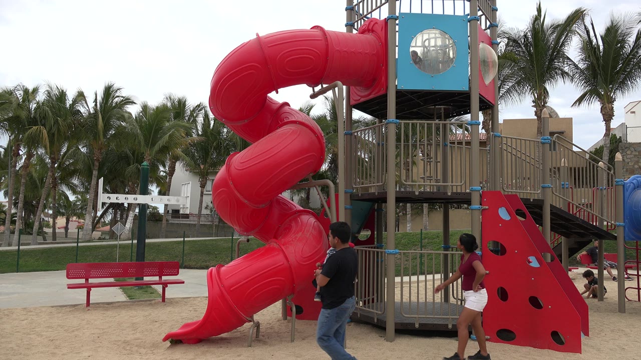 Two adults and a child move across the sandy playground beside the red slide and blue platform in San Jose Del Cabo, Mexico.