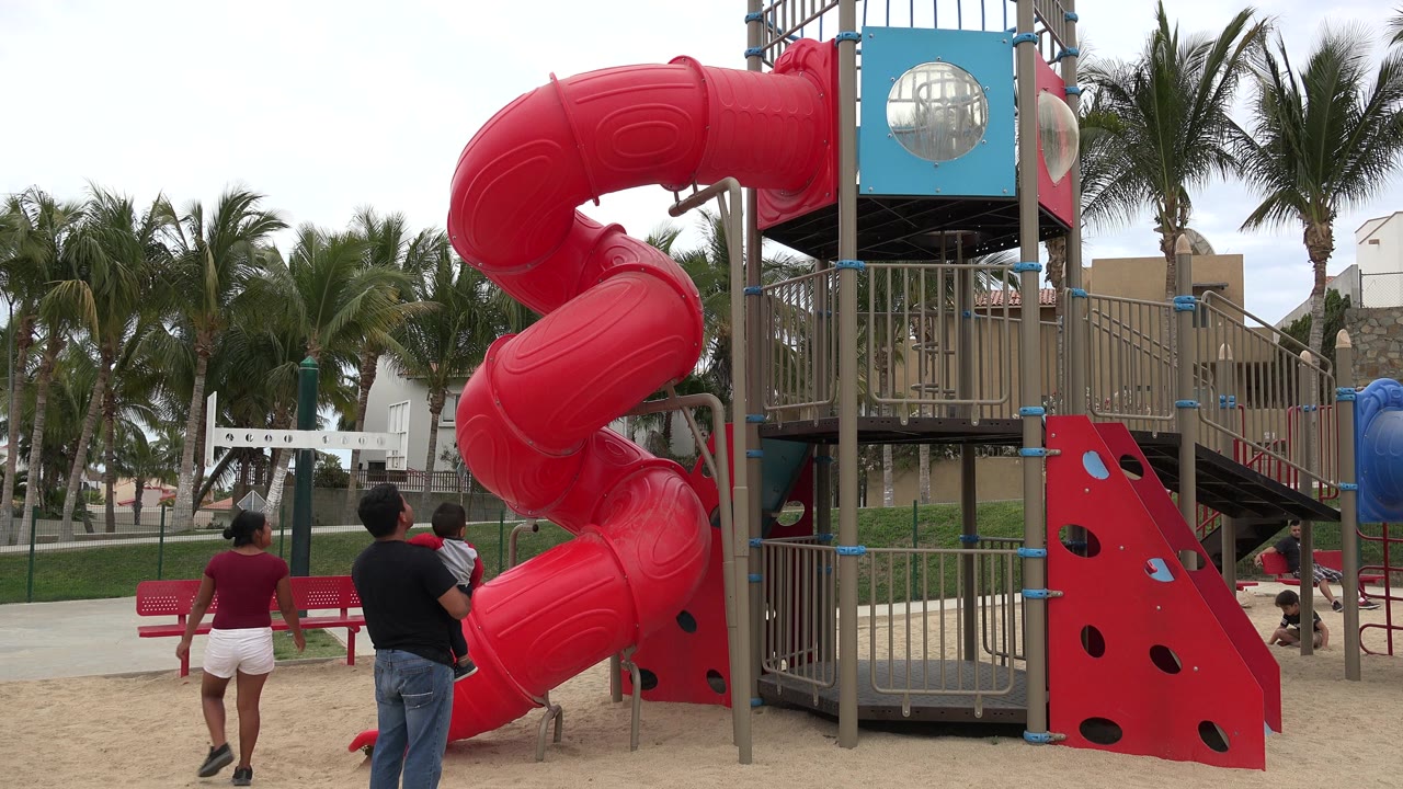 In San Jose Del Cabo, Mexico, adults and a child stand near a large red spiral slide in a sandy playground with palm trees behind it.