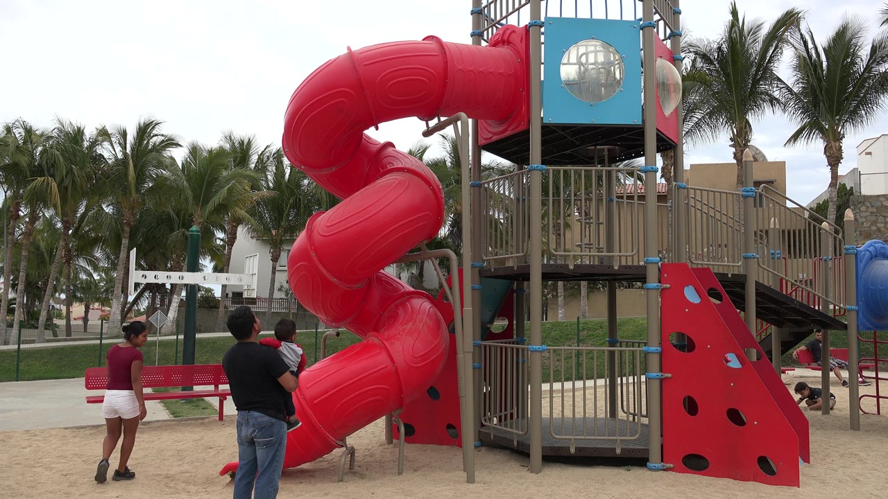 Woman walks away as an adult holds a child near the red slide.