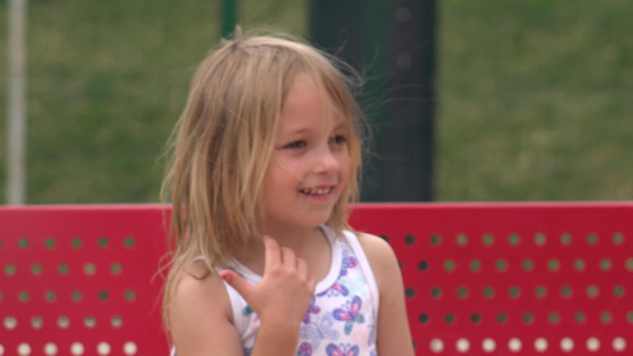 A young blonde girl in a white butterfly-print top smiles beside a red perforated bench in San Jose del Cabo, Mexico, with grass blurred behind her.