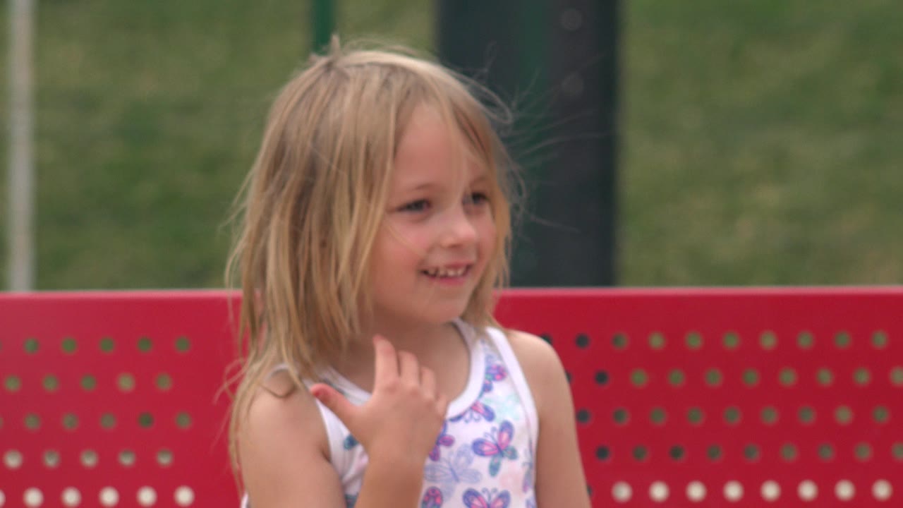 The girl smiles with her hand lifted near her chest, framed against the red bench and soft green background in San Jose del Cabo, Mexico.