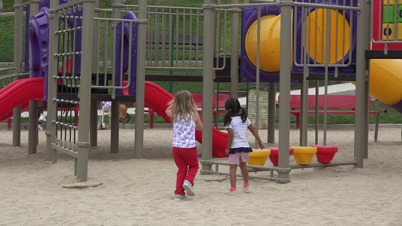 Two young girls walk away from the camera toward gray playground rails and a red slide in San Jose del Cabo, Mexico.