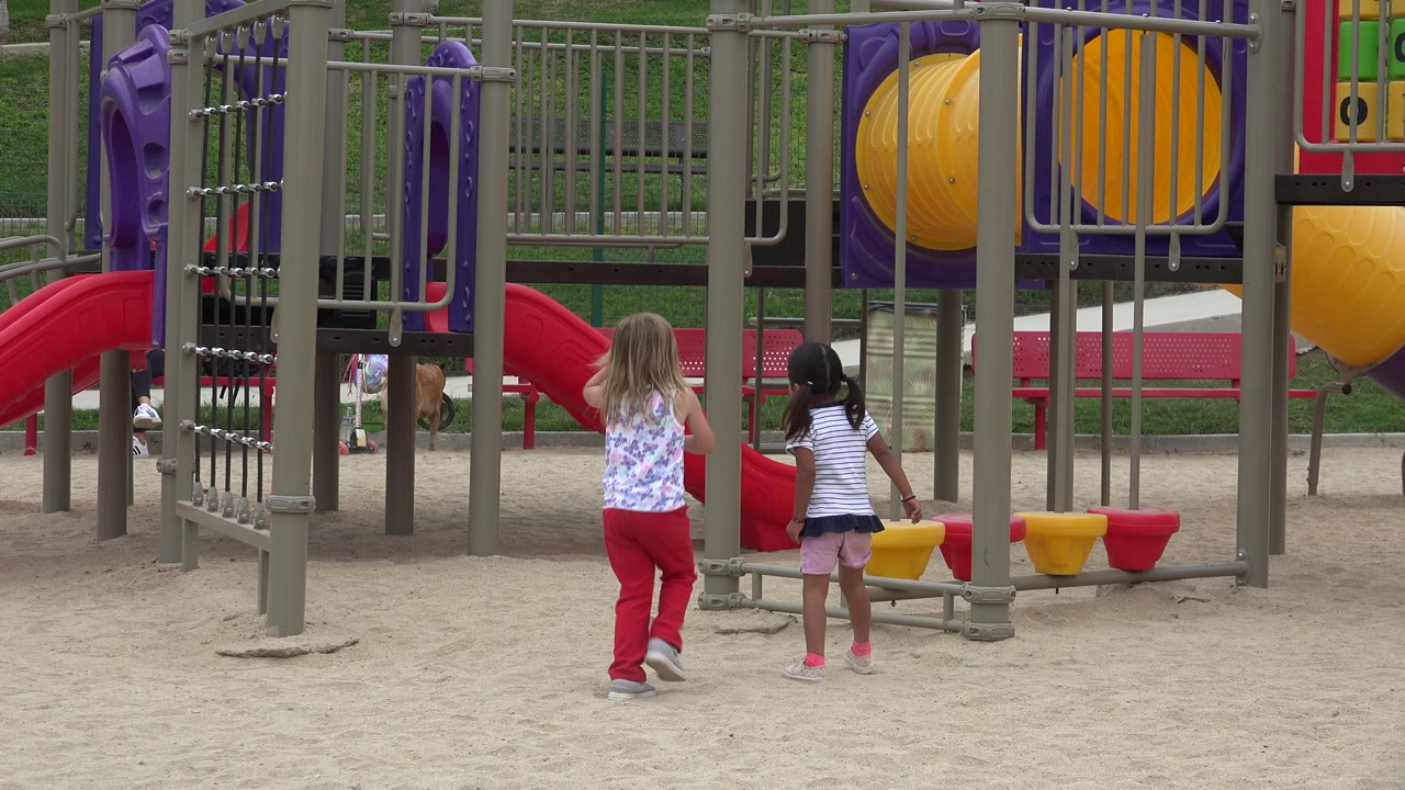 The girls move closer to the lower bars beside the red slide, with yellow and purple tunnel pieces behind them in San Jose del Cabo, Mexico.