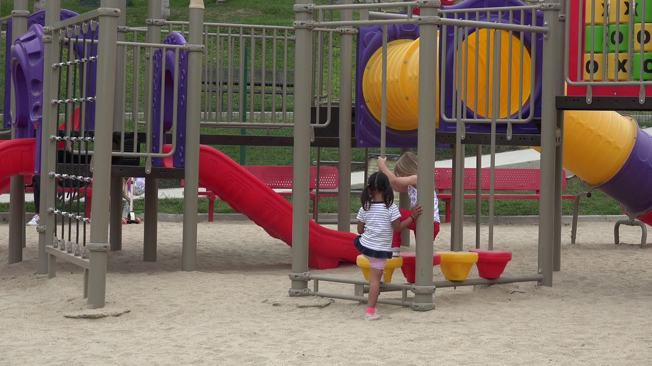 Two small children stand near yellow stepping seats on a sandy playground in San Jose Del Cabo, Mexico, with a red slide, gray bars, and purple equipment behind them.