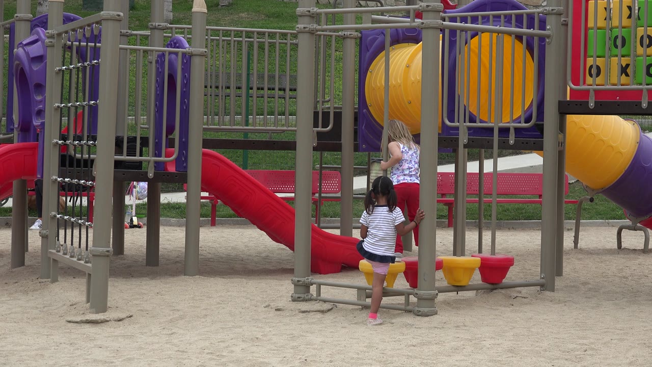 Two children stand by gray playground posts and yellow foot pods.