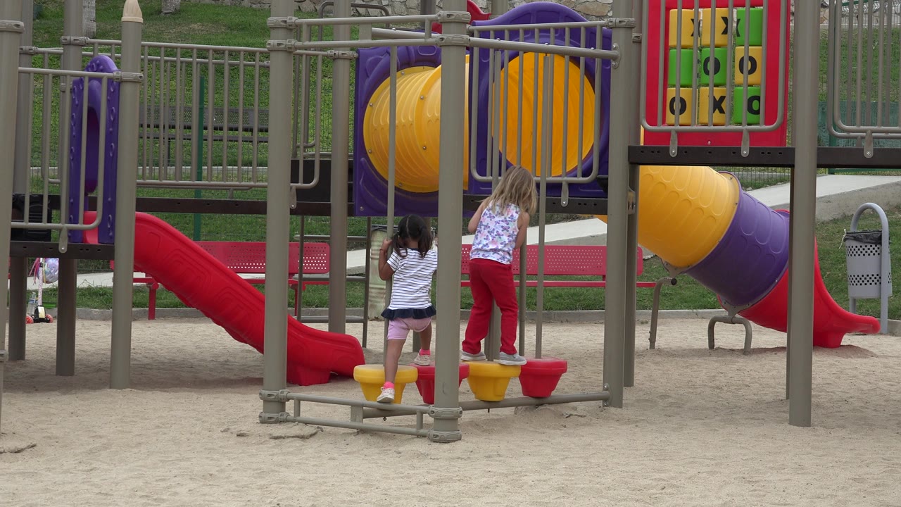 Two children stand on a playground near red slides and yellow stepping pods.
