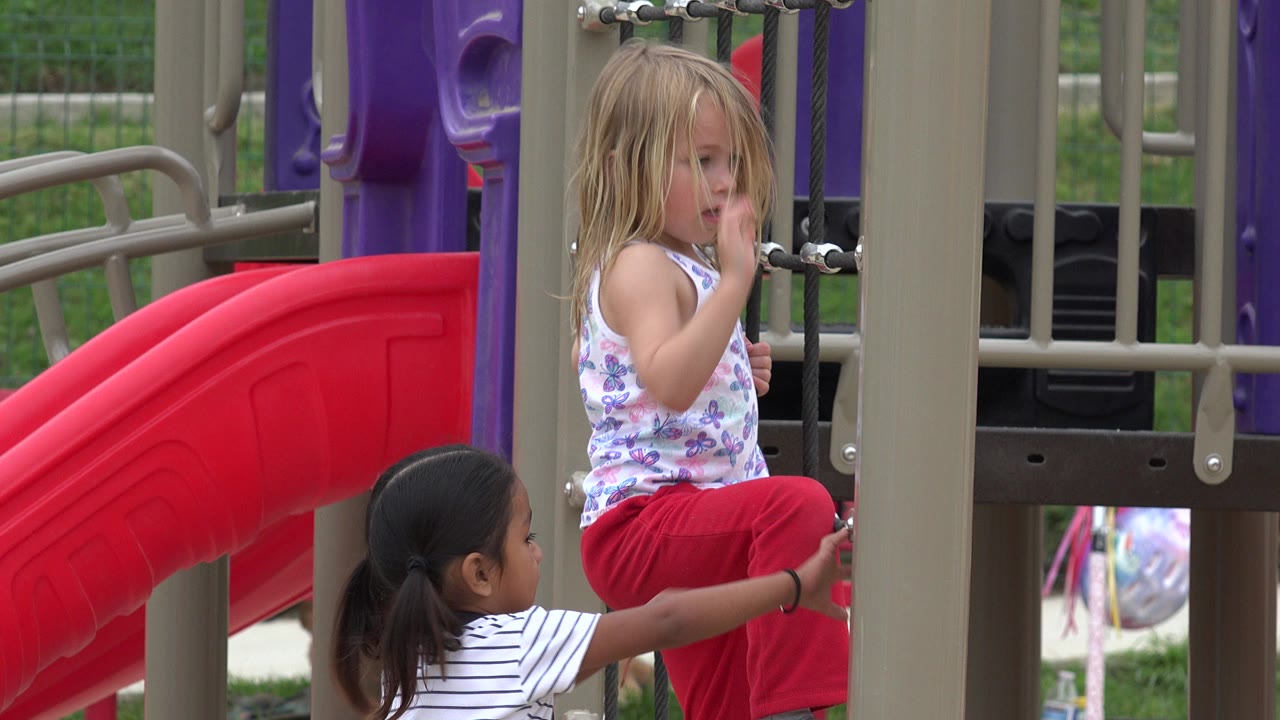 Two young girls play on a colorful playground structure in San Jose Del Cabo, Mexico, with one seated near black ropes and a red slide behind her.