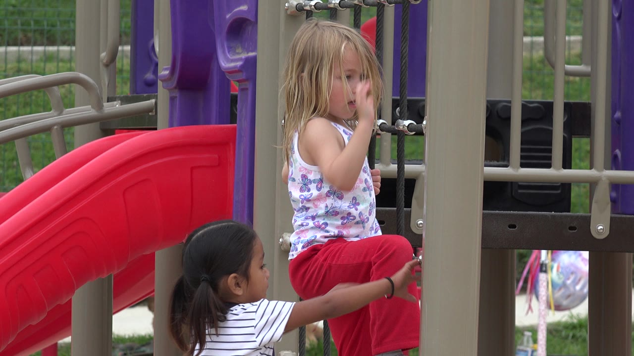 The two girls remain near the rope section of the playground in San Jose Del Cabo, Mexico, with one child seated higher and the other reaching toward the ropes.