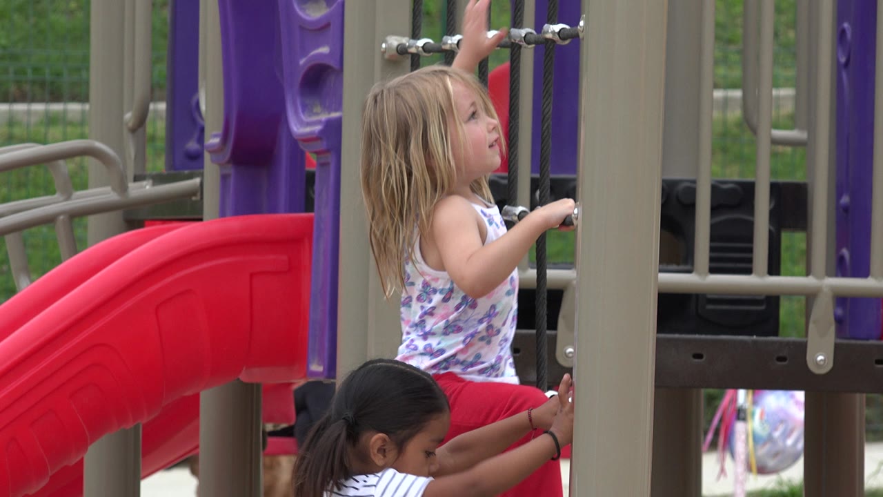 Blonde girl reaches up on playground ropes while another girl stands below.