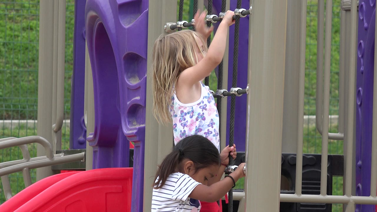 A blonde child reaches up on the rope climber while another girl holds a lower rope near tan playground posts in San Jose Del Cabo, Mexico.