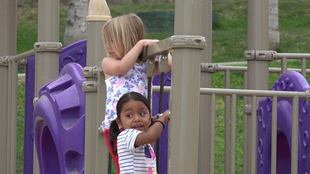 One girl looks toward camera while another leans over a tan playground rail in San Jose Del Cabo, Mexico.