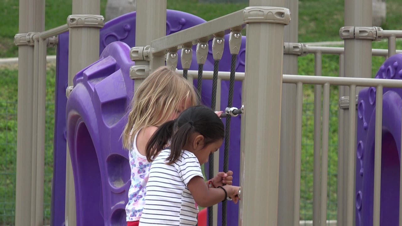The girls remain near the upper part of the black playground net, partially screened by beige posts and purple plastic panels in San Jose Del Cabo, Mexico.