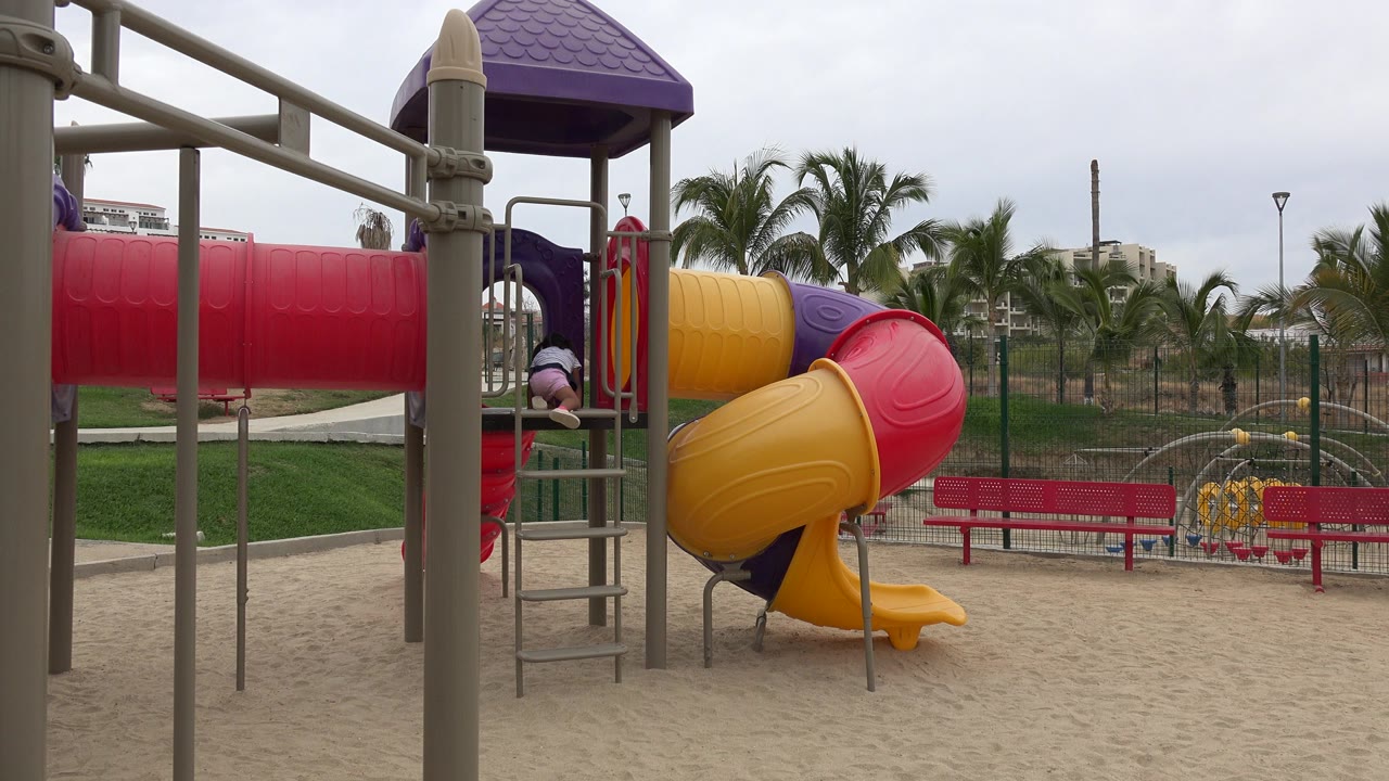 A child in red pants is partly visible on a San Jose del Cabo playground structure, with a red tube slide, yellow spiral slide, gray bars, sand, benches, palms, and cloudy sky.