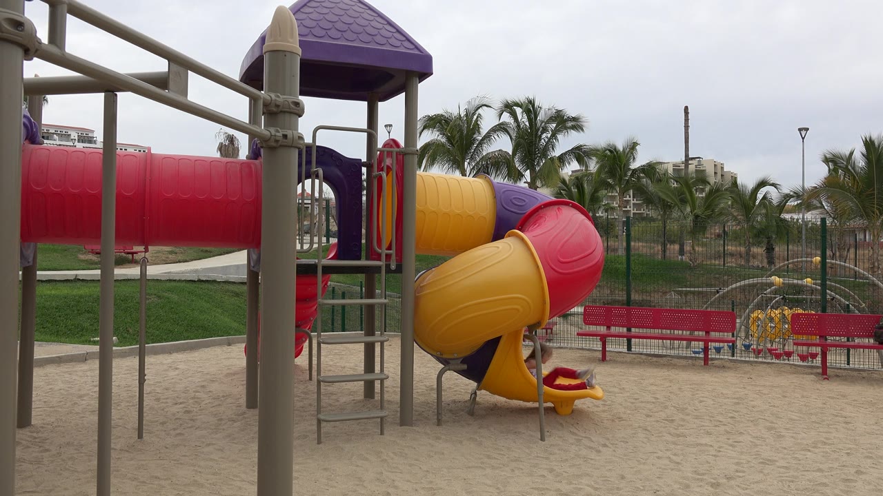Colorful playground slide over sand with red benches and palm trees behind it.
