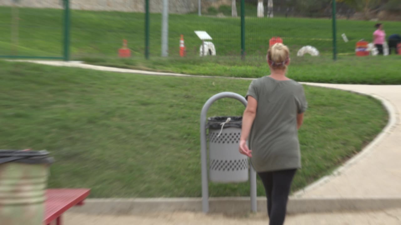 Woman approaches a gray receptacle beside a curving park path.