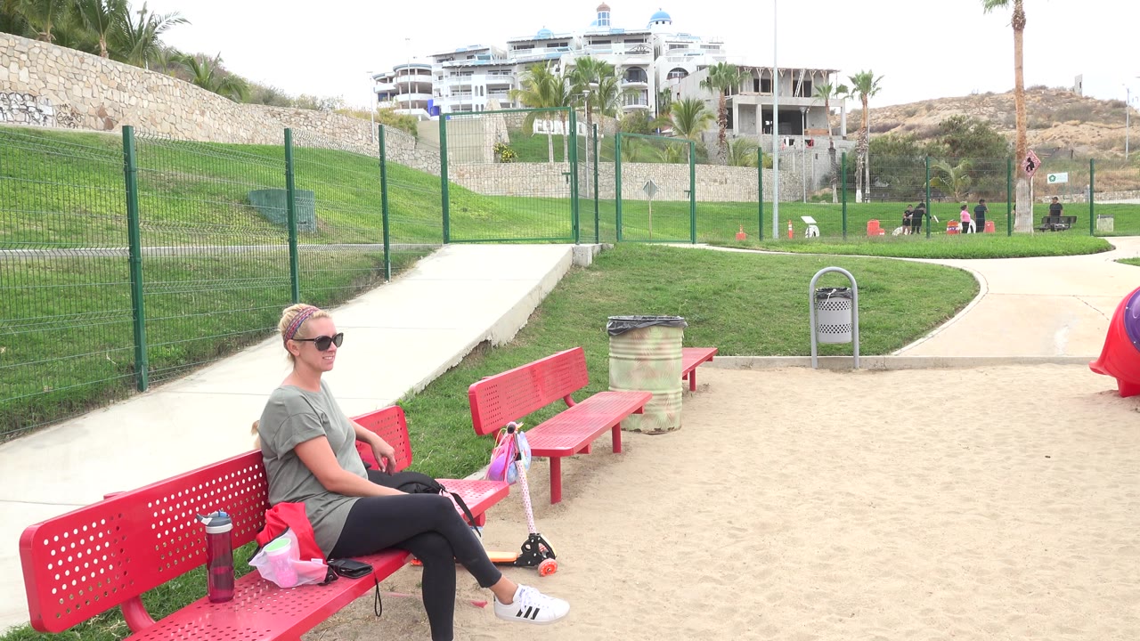 Woman sitting on a red park bench near paths and grass.