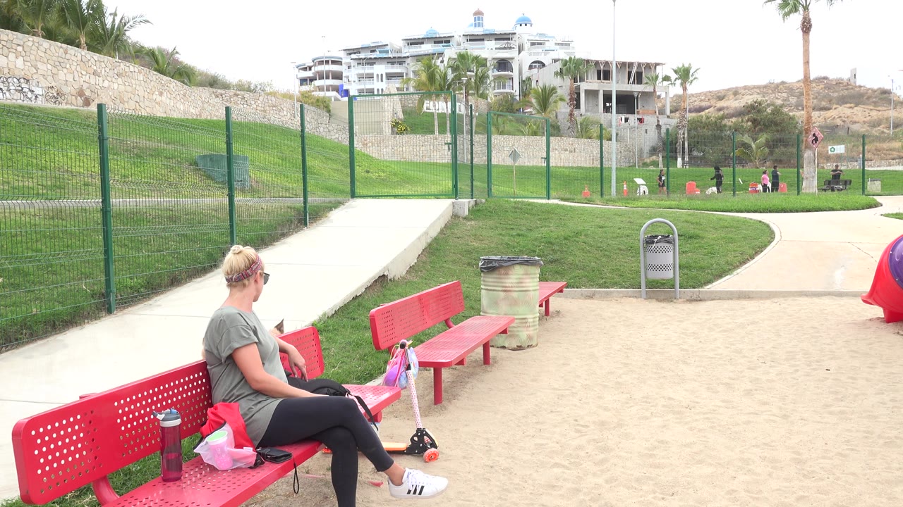 Woman seated on a red park bench beside bags and a scooter.