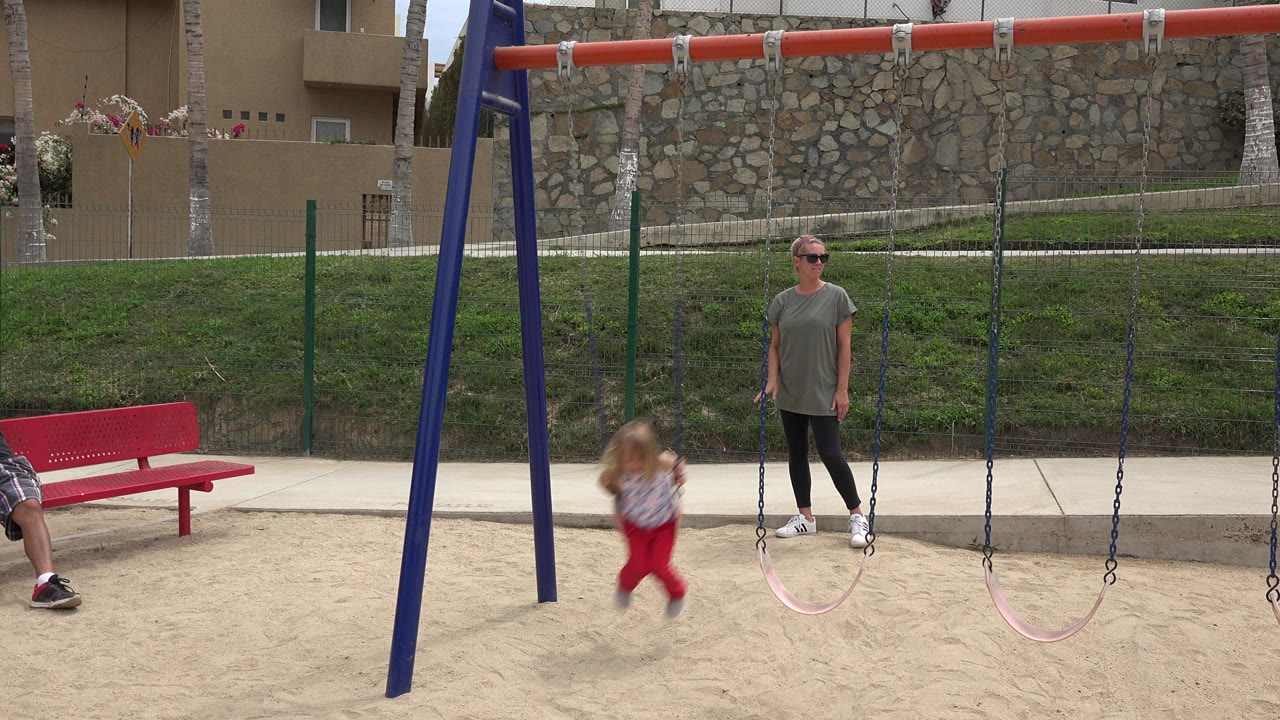Woman beside swing set while a little girl swings forward in a park.