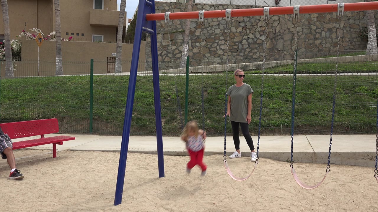 In San Jose del Cabo, Mexico, the little girl swings near the lower part of the arc while the woman waits beside adjacent empty swings, with the red bench partly visible at left.