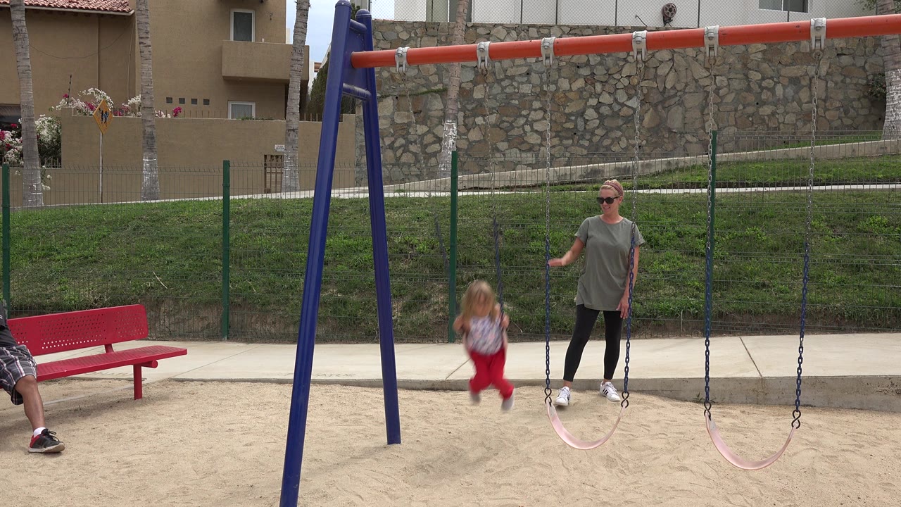 Child on a swing with an adult woman standing nearby at a sandy playground.