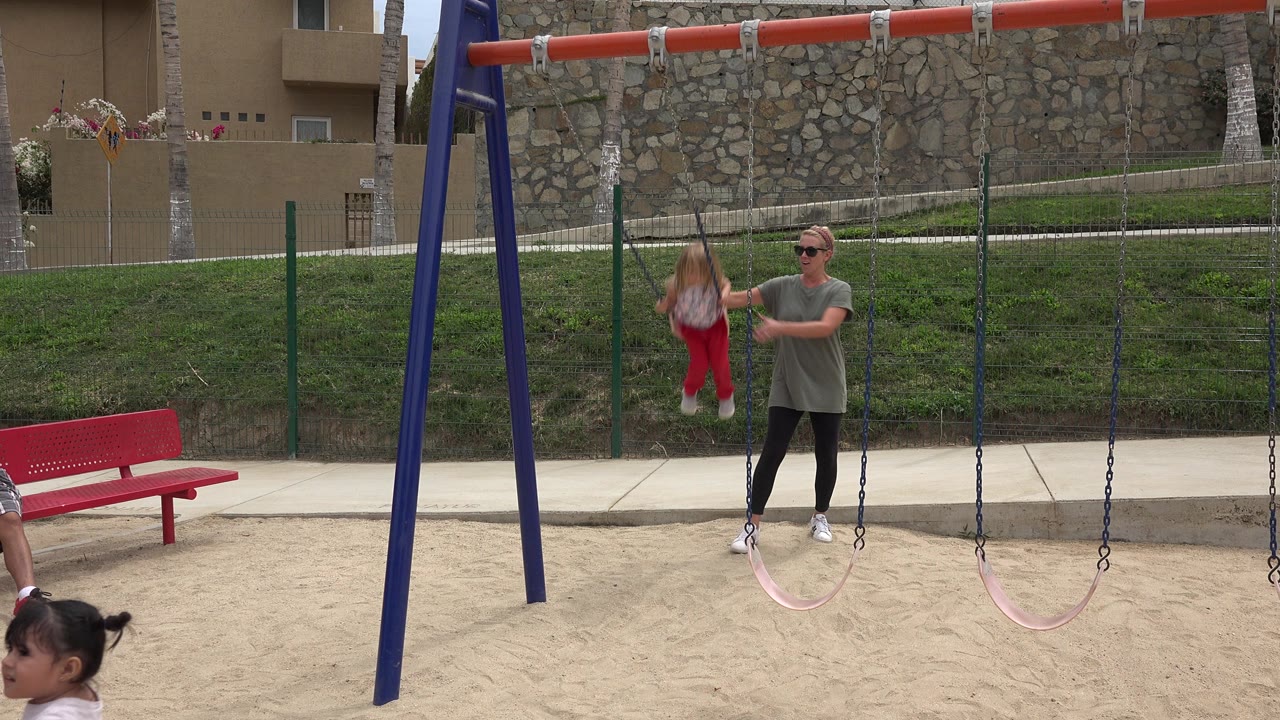Mother in sunglasses stands near a young girl on a playground swing.