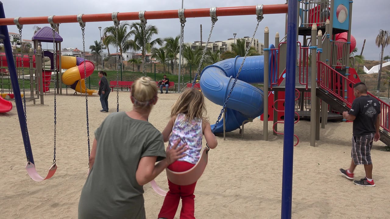 Woman pushes a young girl on a playground swing.