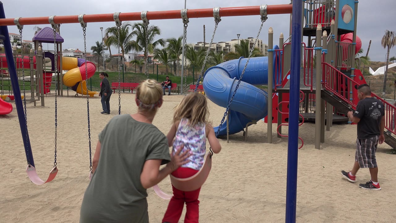 The woman leans closer behind the seated girl on the swing in San Jose Del Cabo, Mexico, while a man stands near the blue slide and empty swing chains hang in front.