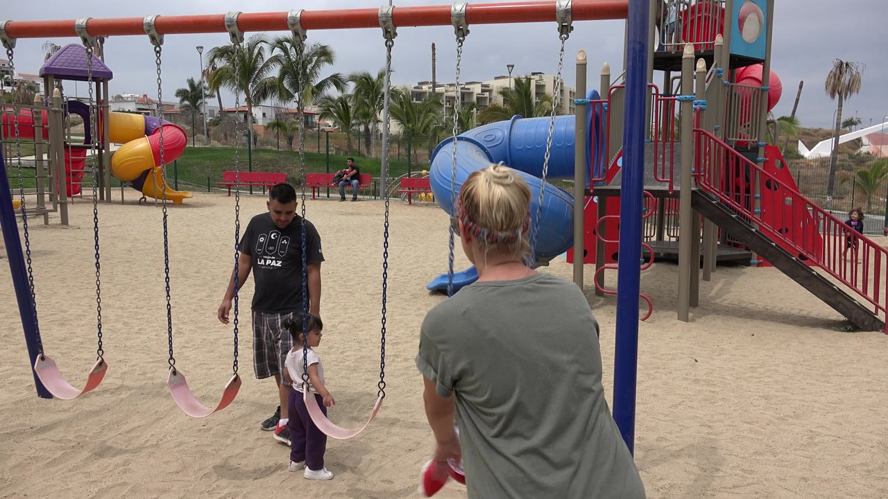 At a San Jose Del Cabo public playground, an adult man stands beside a small child near pink swing seats while a blonde adult is seen from behind in the foreground.